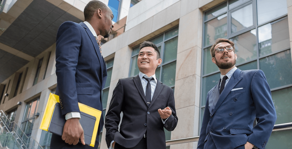 Three businessmen in suits converse outside a modern office building, one holding documents and a tablet.
