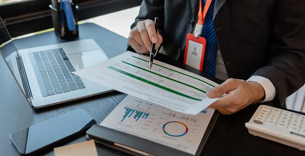 Businessperson in a suit reviews financial documents with charts, using a pen beside a laptop on a desk.